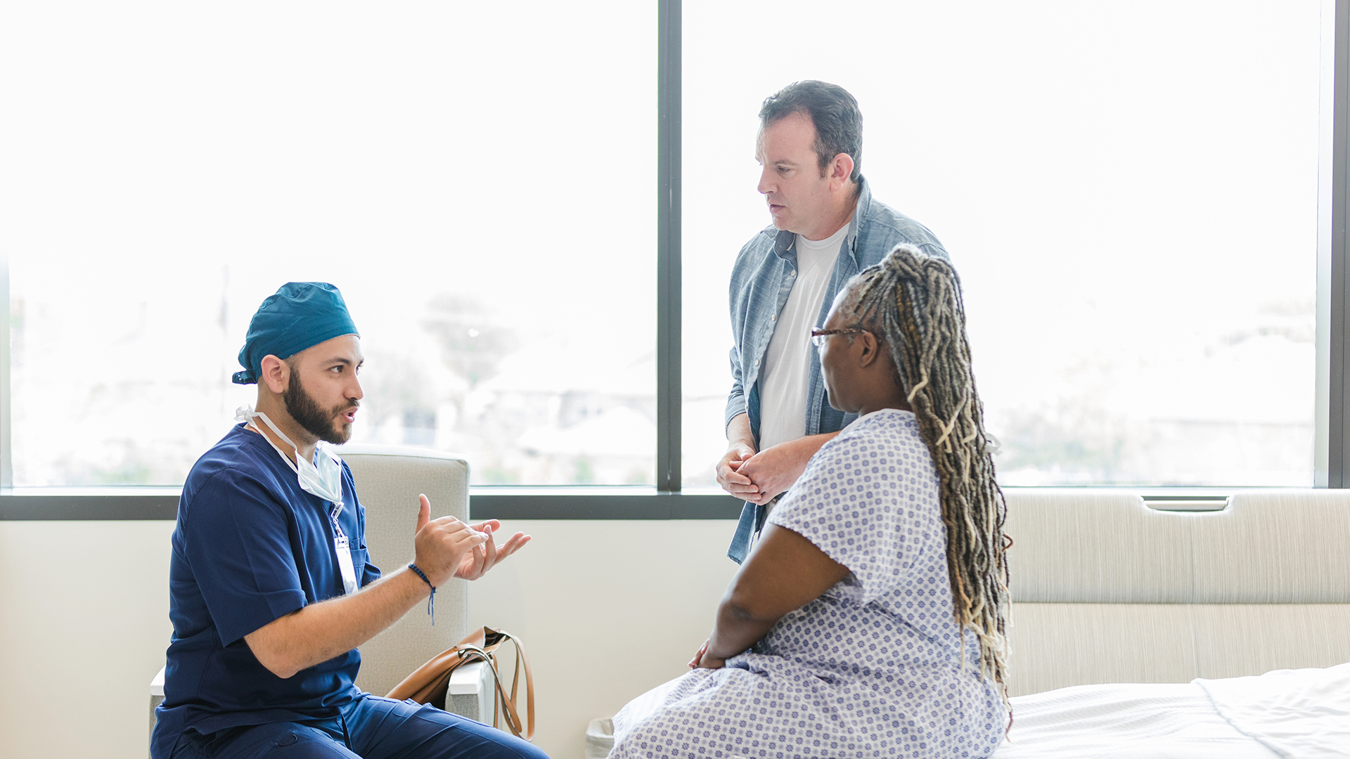 A patient male doctor gives step-by-step detail of a female patient's surgery as the patient and her husband listen.