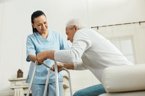 Professional caregiver. Joyful nice pleasant woman smiling and looking at the elderly man while helping him to walkhelping elderly patient stand up using walker