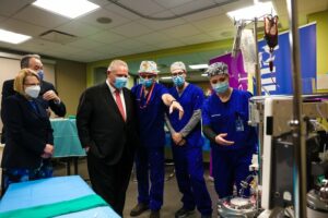Michener students and a professor in masks and scrub caps stand beside Premier Doug Ford and Health Minister Sylvia Jones in a simulated operating room