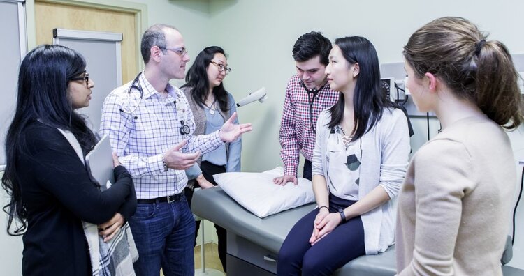 Several students in a doctor's office with a patient bed looking at an instructor speaking