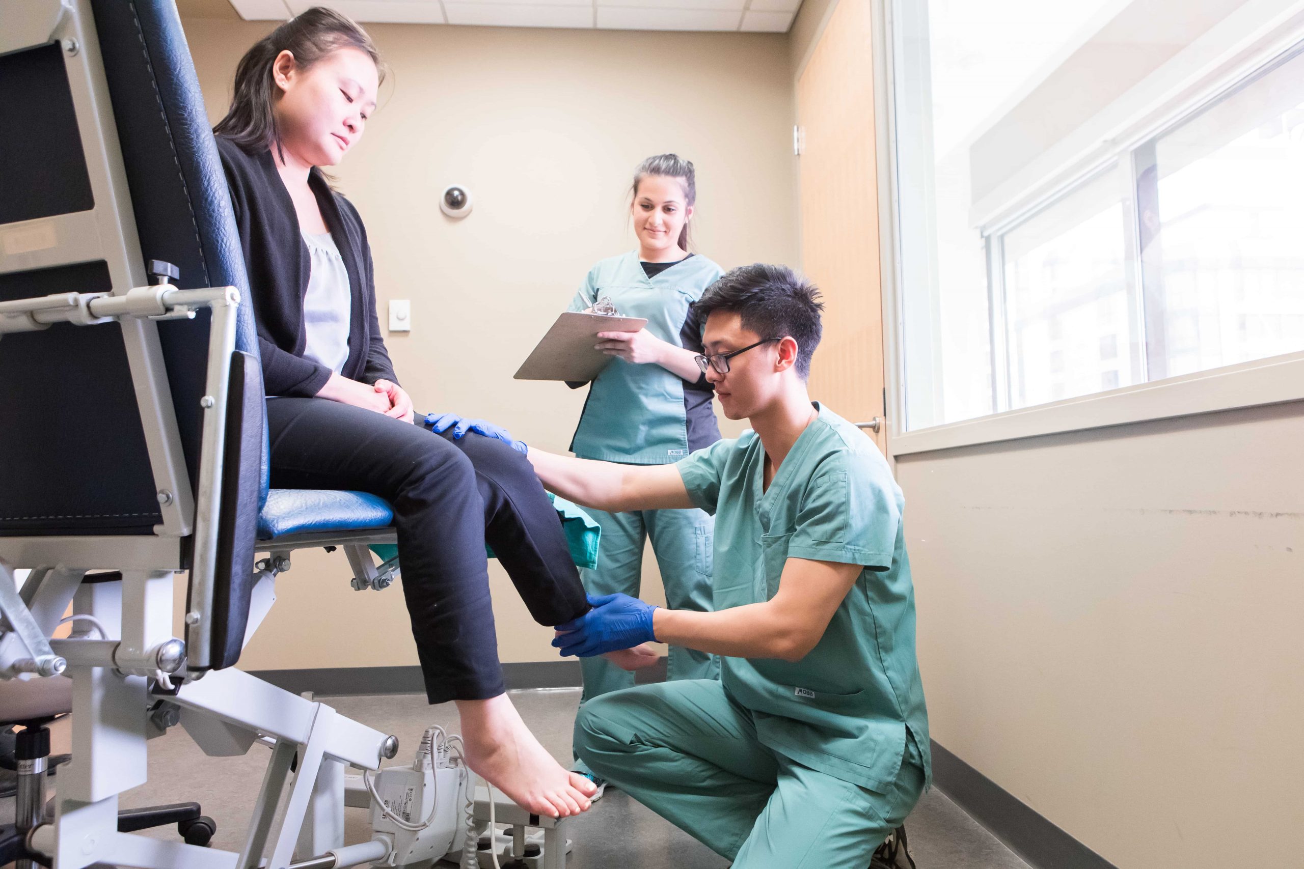 A Chiropody practitioner bend's a woman's knee while a trainee looks on.