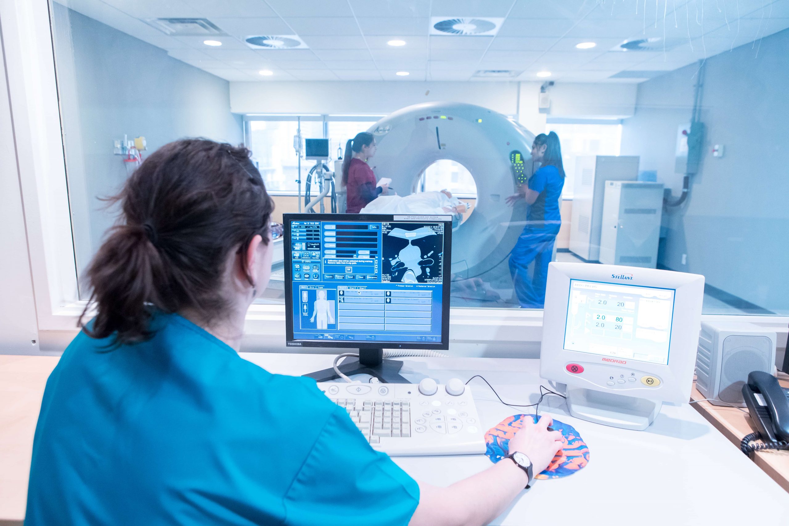 A Michener student in scrubs sits behind a computer while two nurses prep a patient for an MRI