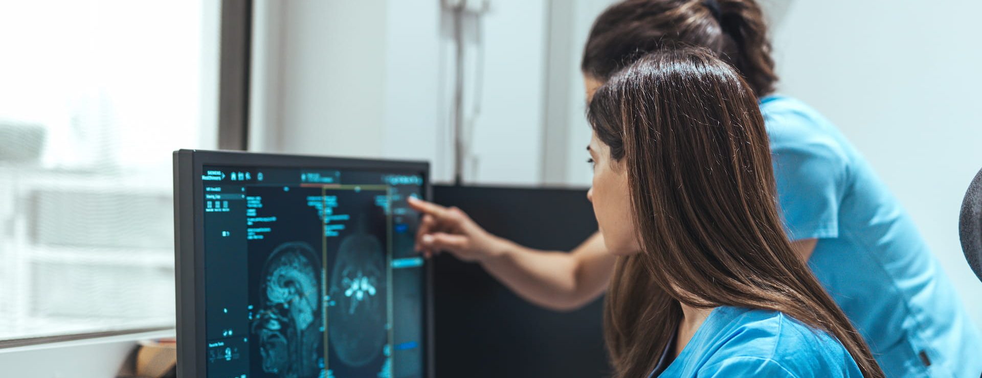 Two healthcare workers in scrubs examine brain scan images on a computer monitor in a medical office.