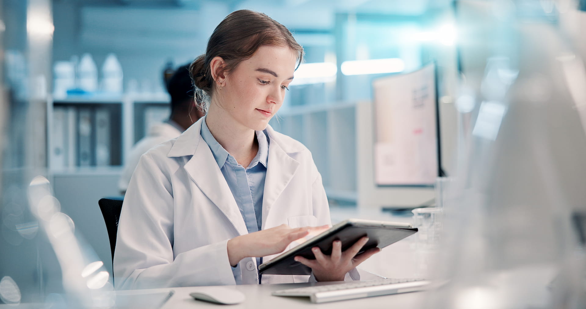 A scientist in a lab coat reviews data on a tablet in a modern laboratory setting.