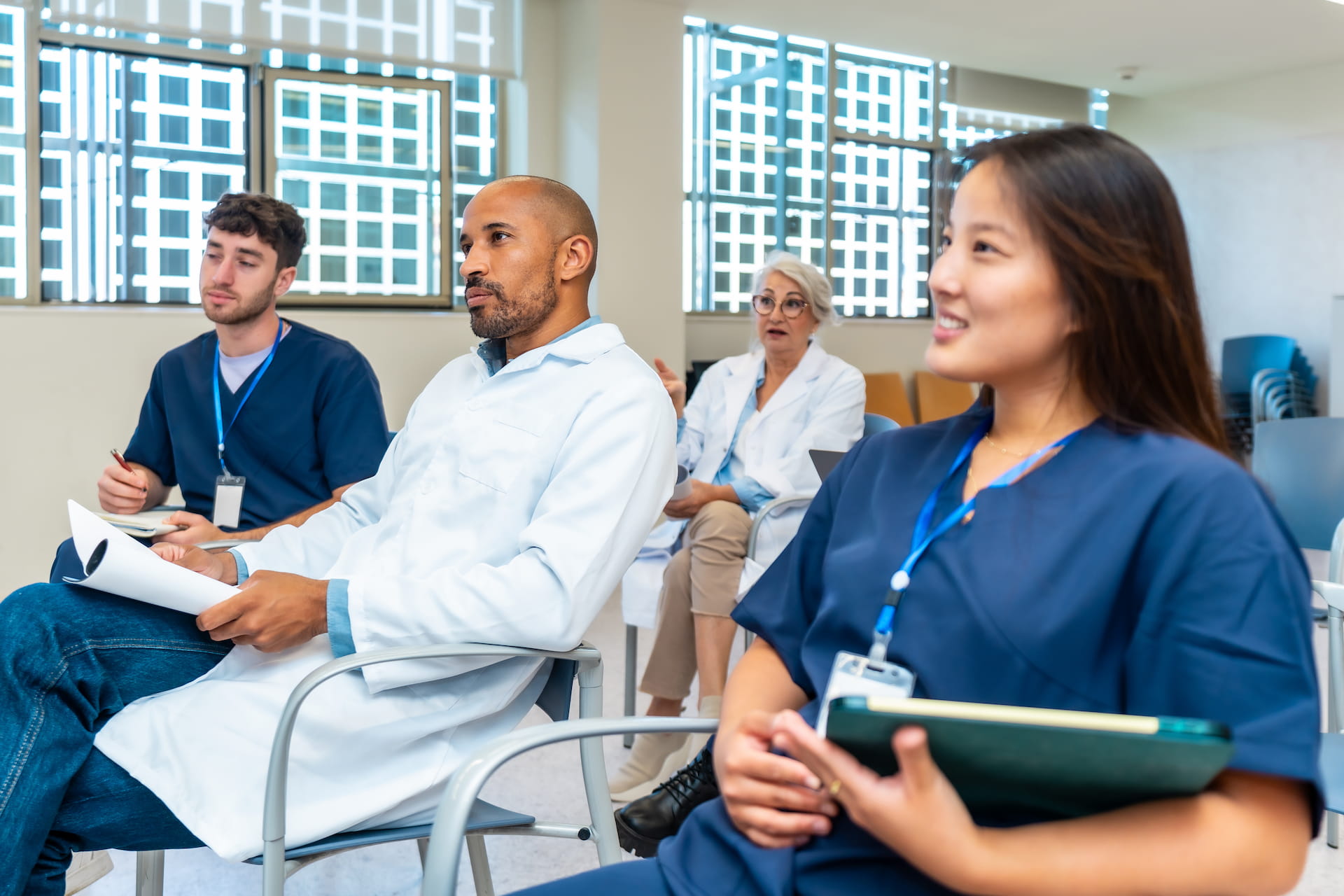 A group of medical professionals in scrubs and lab coats attend a lecture or meeting in a bright room.