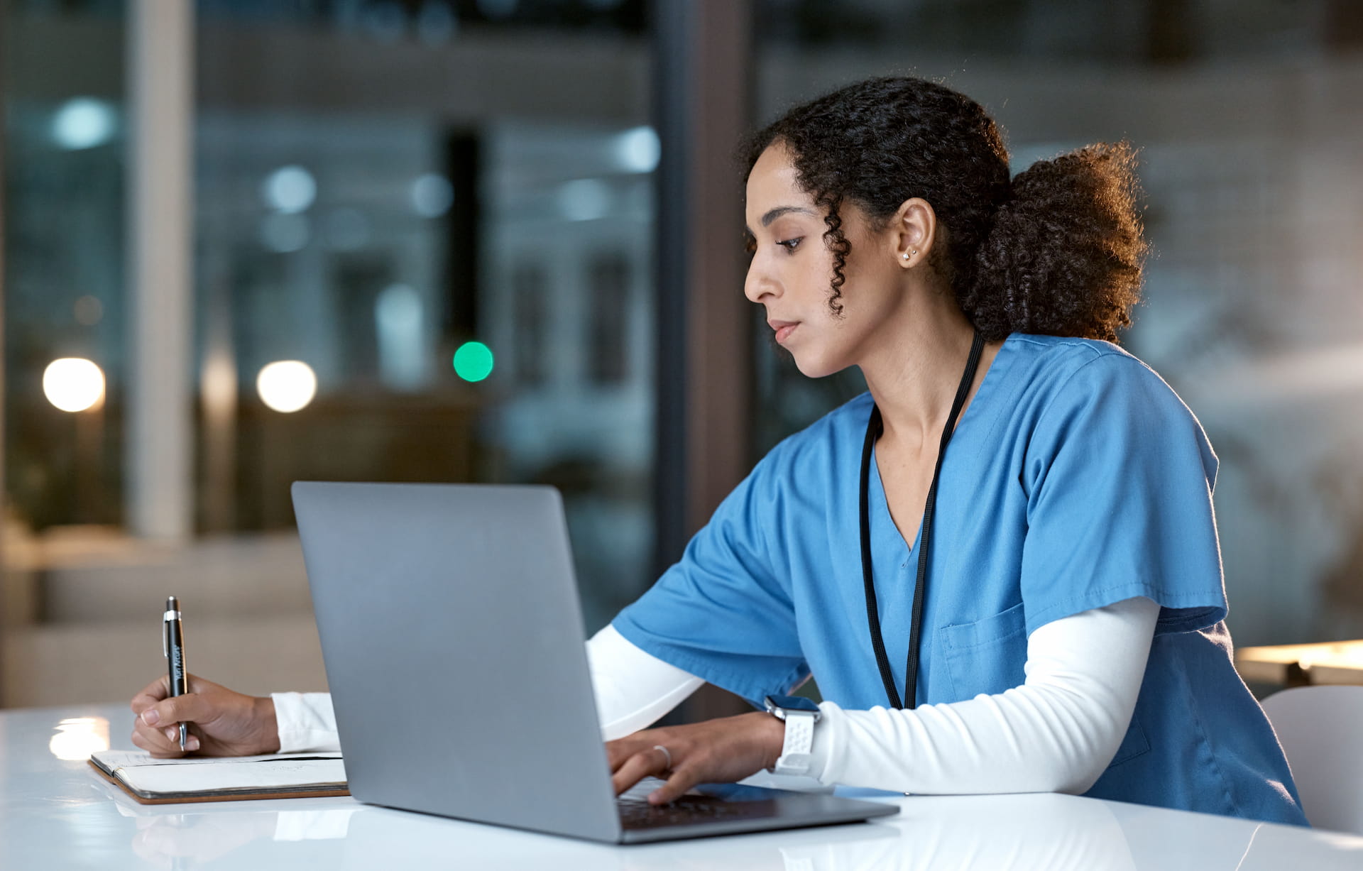 A woman in medical scrubs works at a laptop and writes notes on paper in a brightly lit office setting.