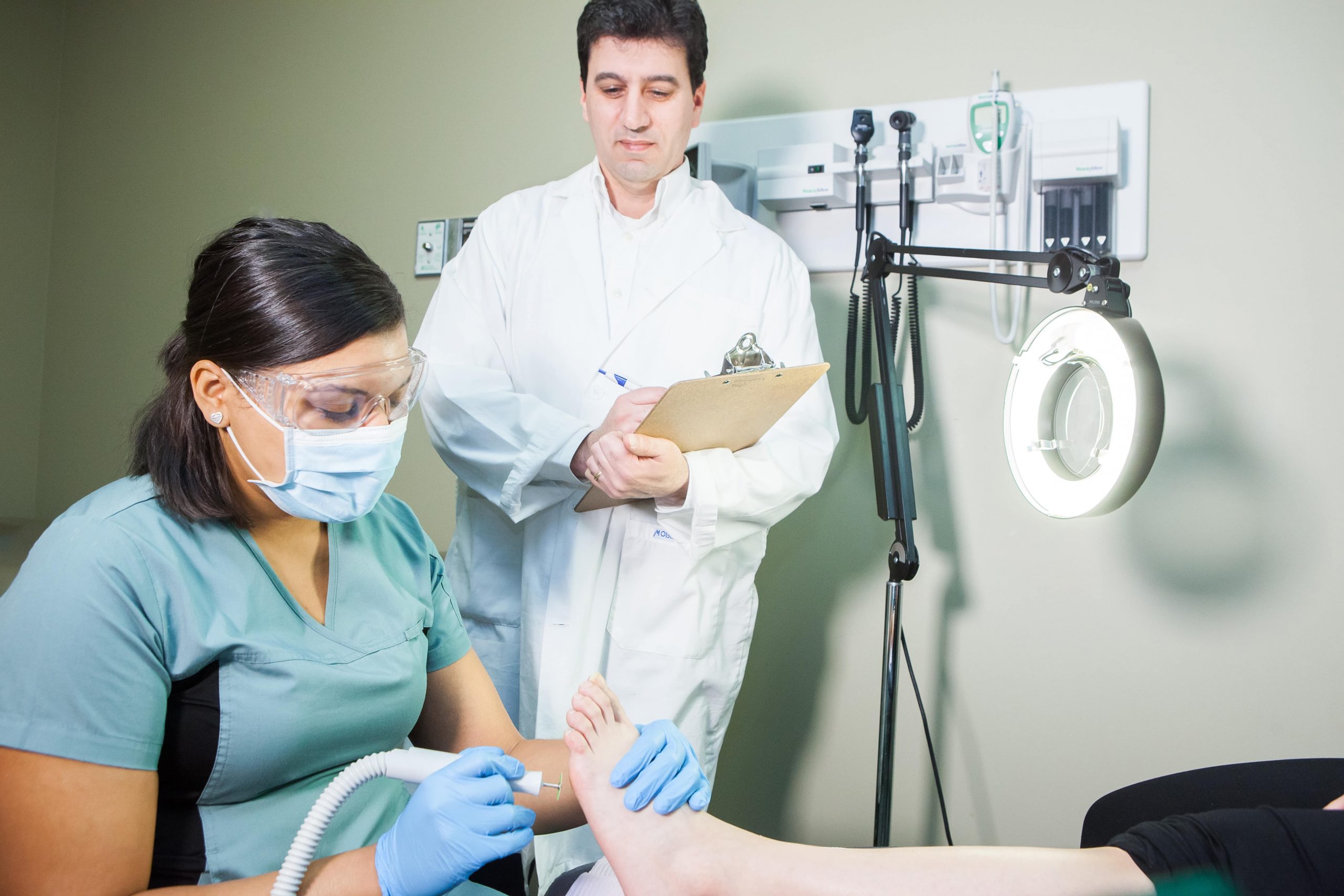 Healthcare worker treats a patient's foot while a doctor observes and takes notes in a medical office.