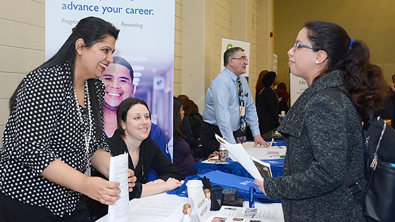 Four people interact at a career fair booth, exchanging resumes and information across a display table.