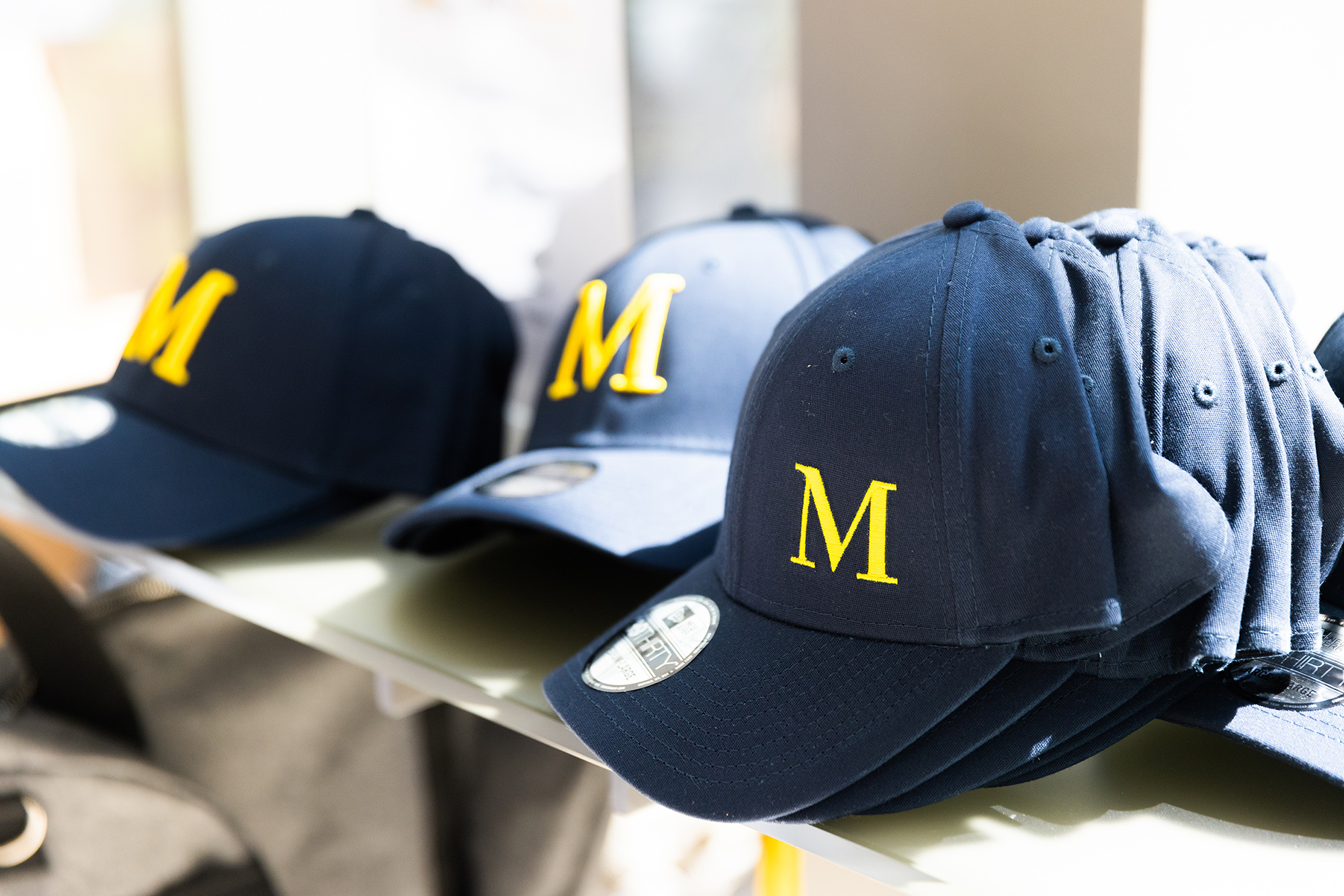 Several navy blue baseball caps with a yellow letter "M" for Michener are stacked on a shelf in bright lighting.