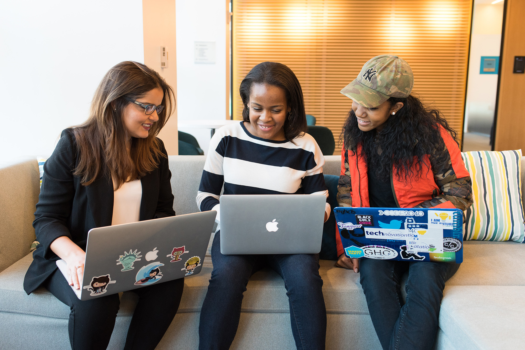Three women sitting on a couch, working together on laptops covered with various stickers.