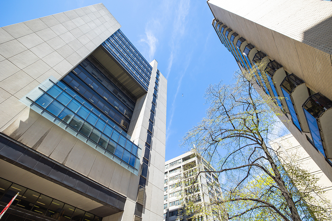 Tall modern office buildings and a tree are seen from a low angle against a bright blue sky with scattered clouds.