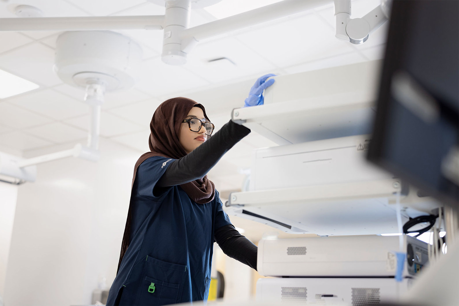 A healthcare worker in scrubs and a hijab organizes medical equipment in a hospital setting.