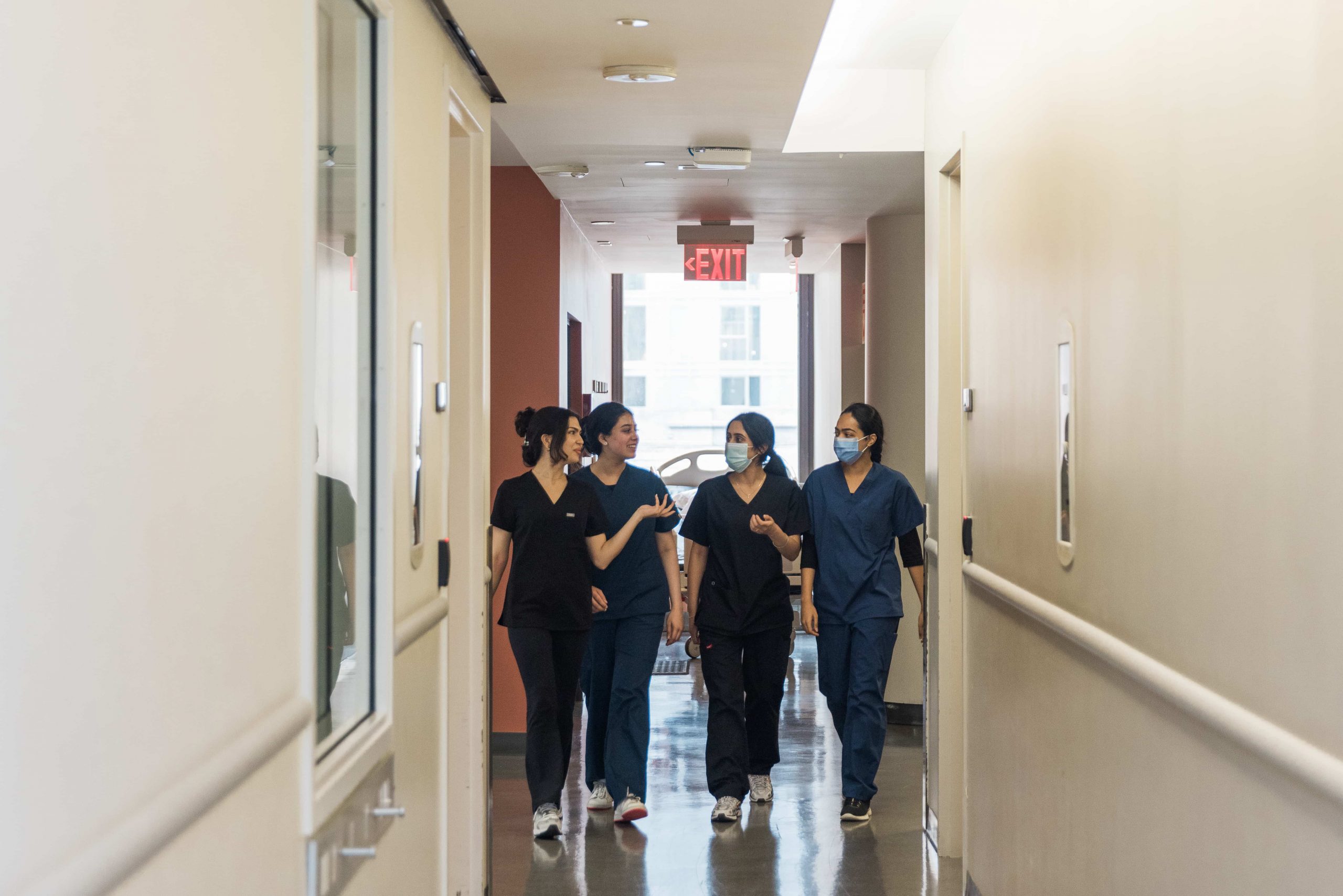 Four healthcare workers in scrubs and masks walk and talk together down a hospital hallway.