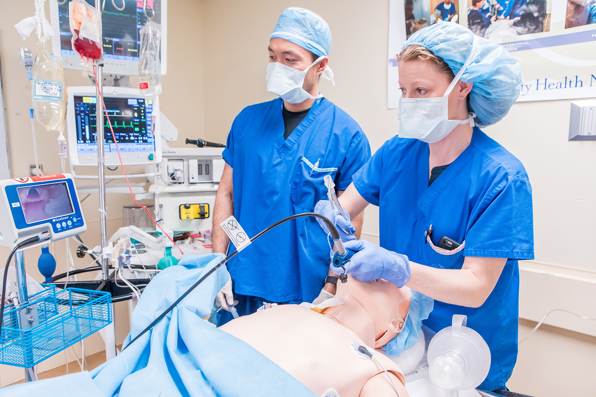 Two medical professionals in scrubs practice intubation on a medical mannequin in a hospital simulation room.