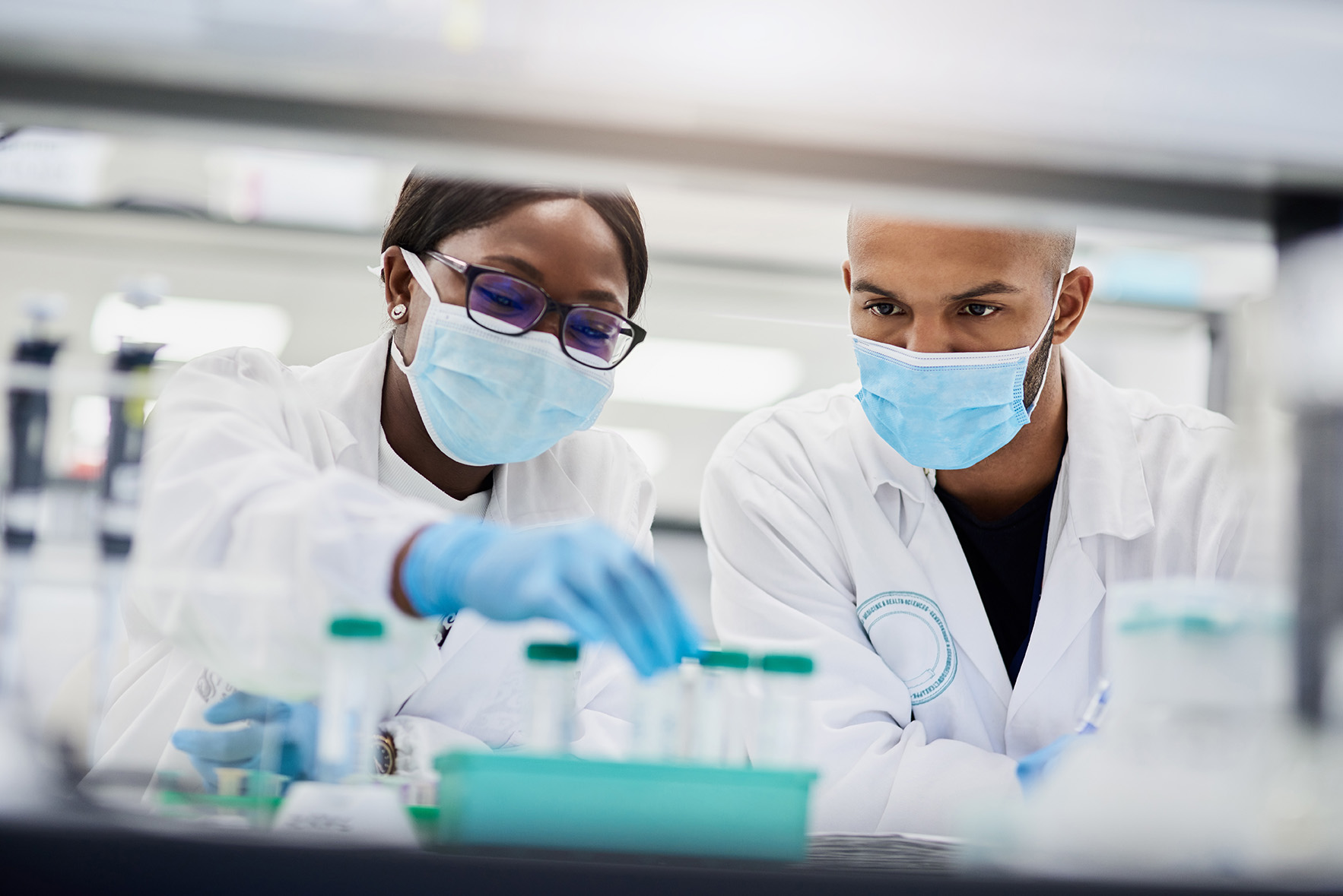 Two scientists wearing lab coats and masks examine samples in test tubes in a laboratory setting.