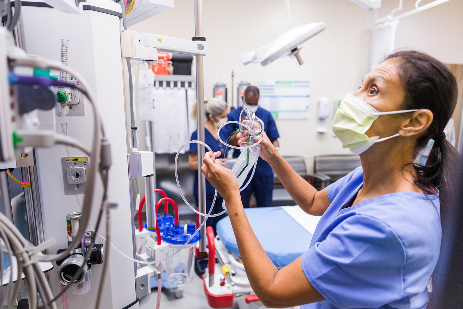A healthcare worker in scrubs and a mask prepares medical equipment in a hospital room.