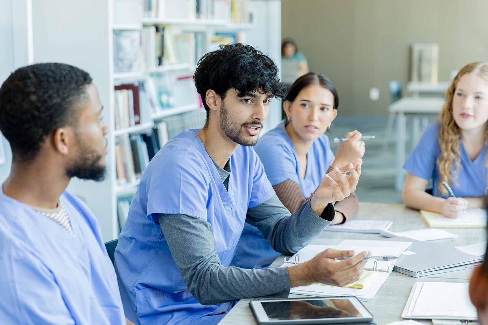 Four medical students in scrubs sit at a table in discussion with notebooks, textbooks, and a tablet in front of them.