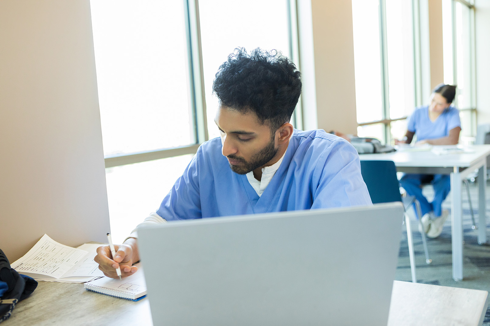 A student in blue scrubs studies at a desk with a laptop and notes, while another person works in the background.