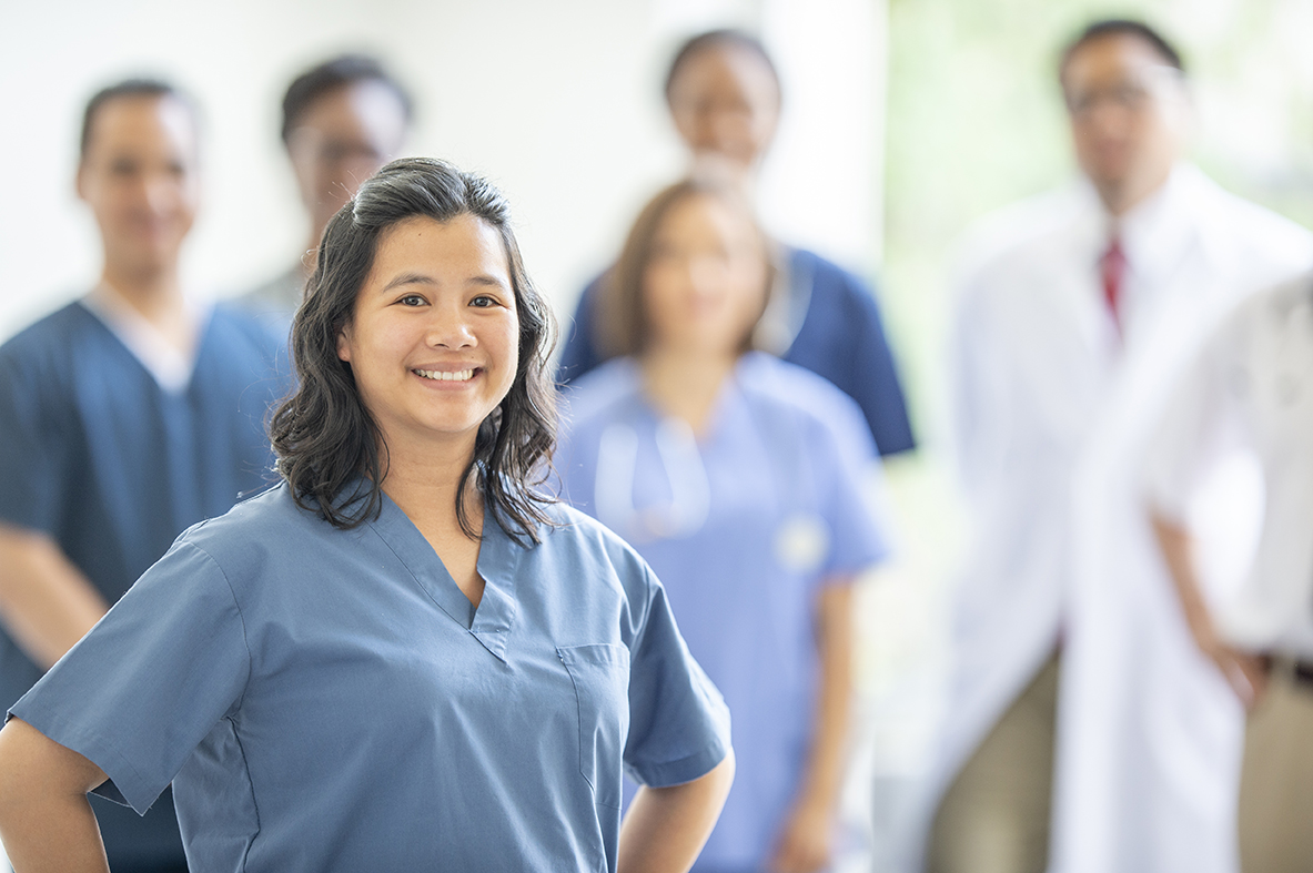 A woman in medical scrubs stands in front of a group of healthcare professionals in a bright setting.