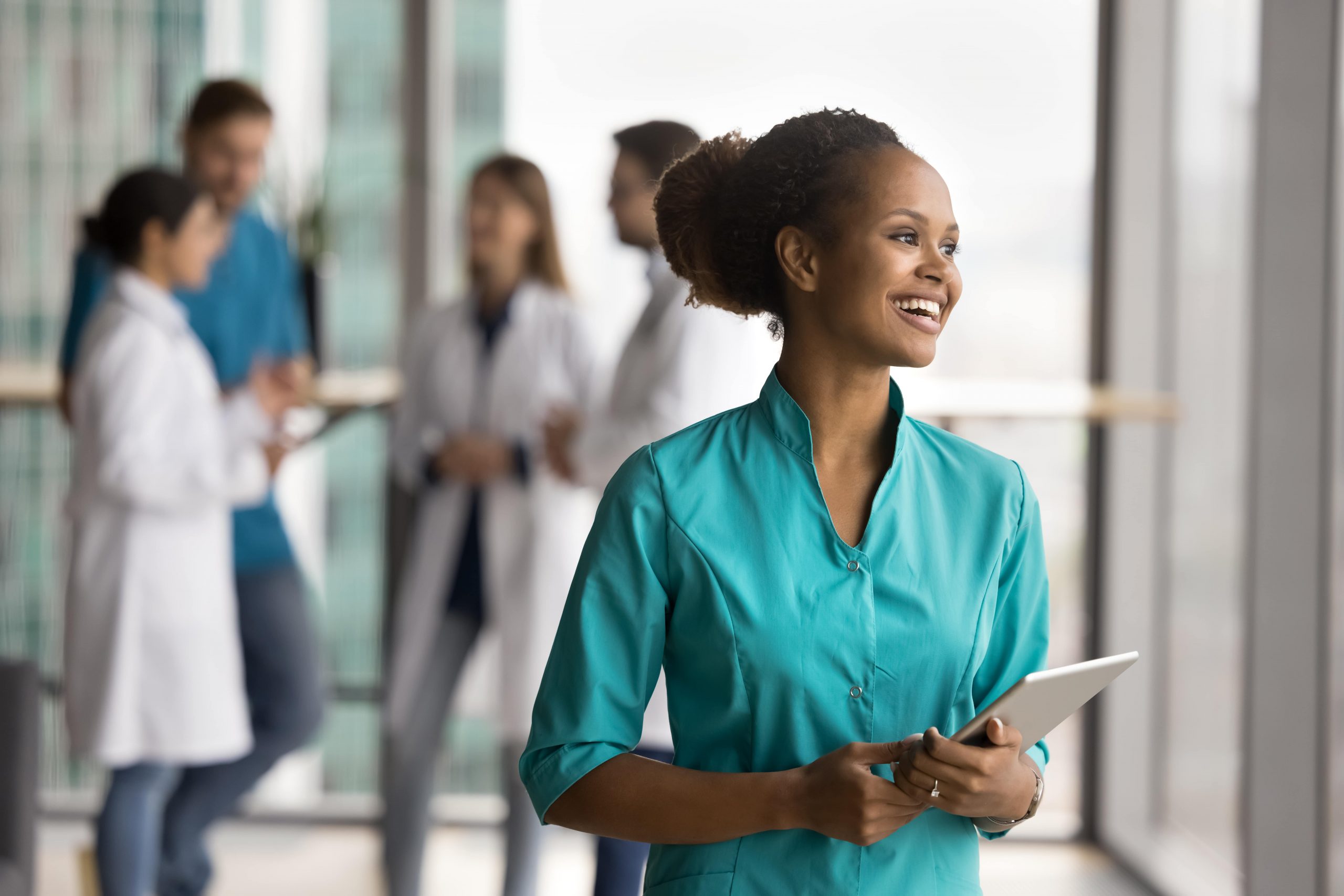 Smiling healthcare worker holding a tablet, with colleagues in lab coats talking in the background of a modern office.
