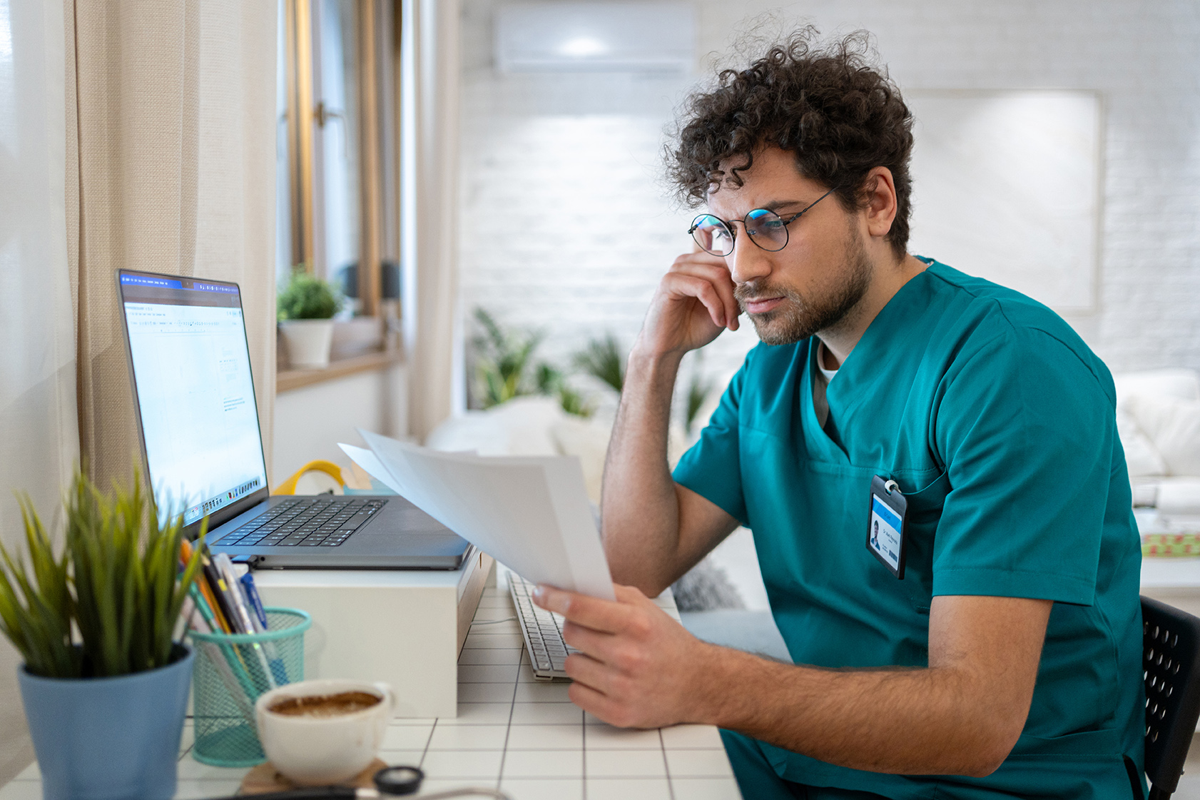 A healthcare worker in scrubs reviews documents at a desk with a laptop and office supplies.