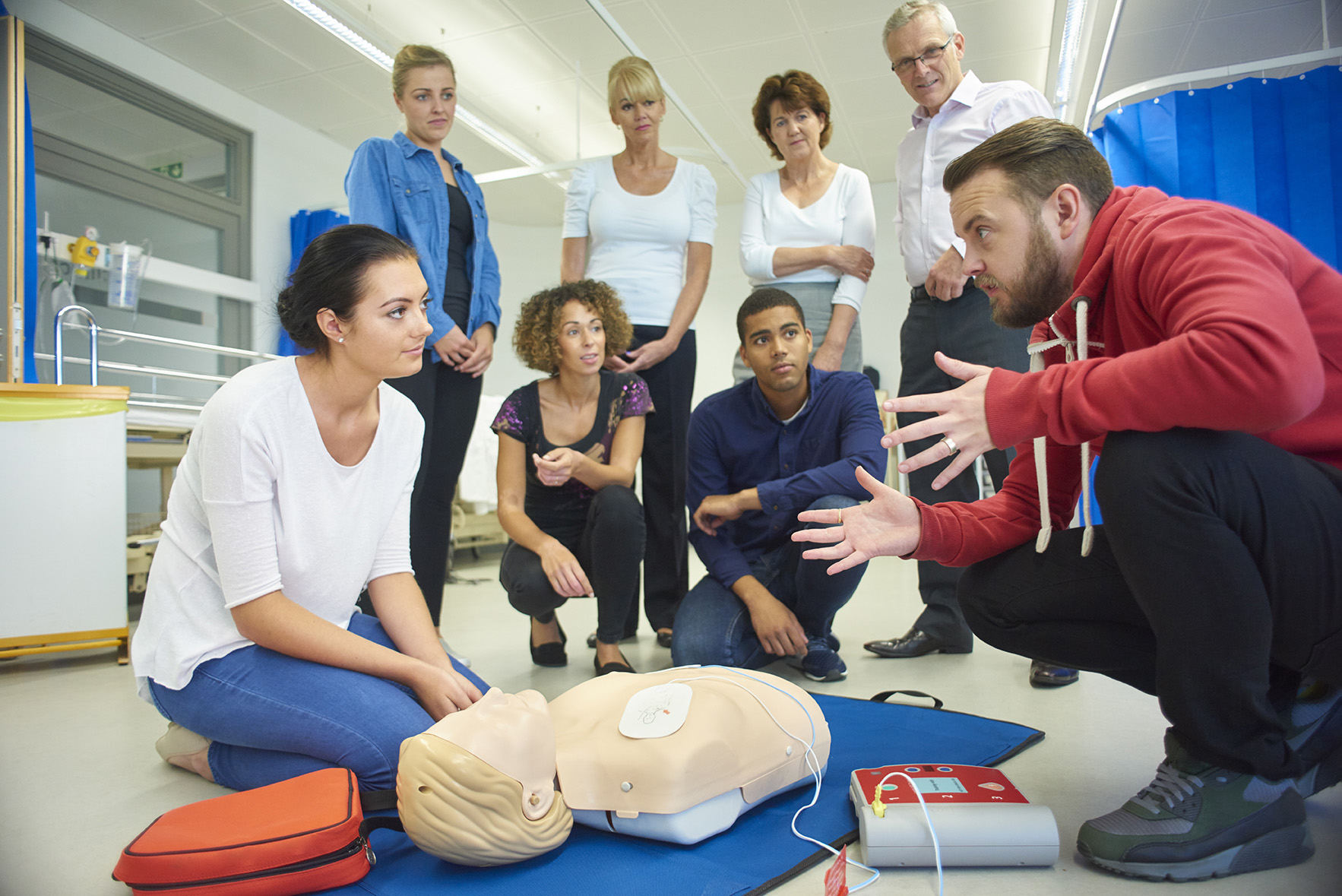 A group of adults watch an instructor demonstrate CPR and AED use on a training manikin in a classroom setting.