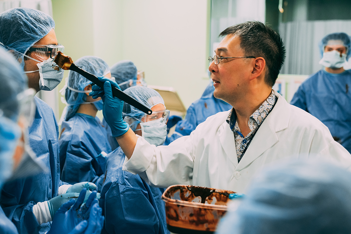 A man in a lab coat applies brown substance to a person's goggles while others in scrubs watch in a medical setting.