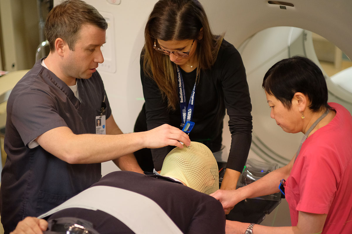 Three medical professionals fit a mesh mask onto a patient’s head for a procedure, possibly in a radiology setting.
