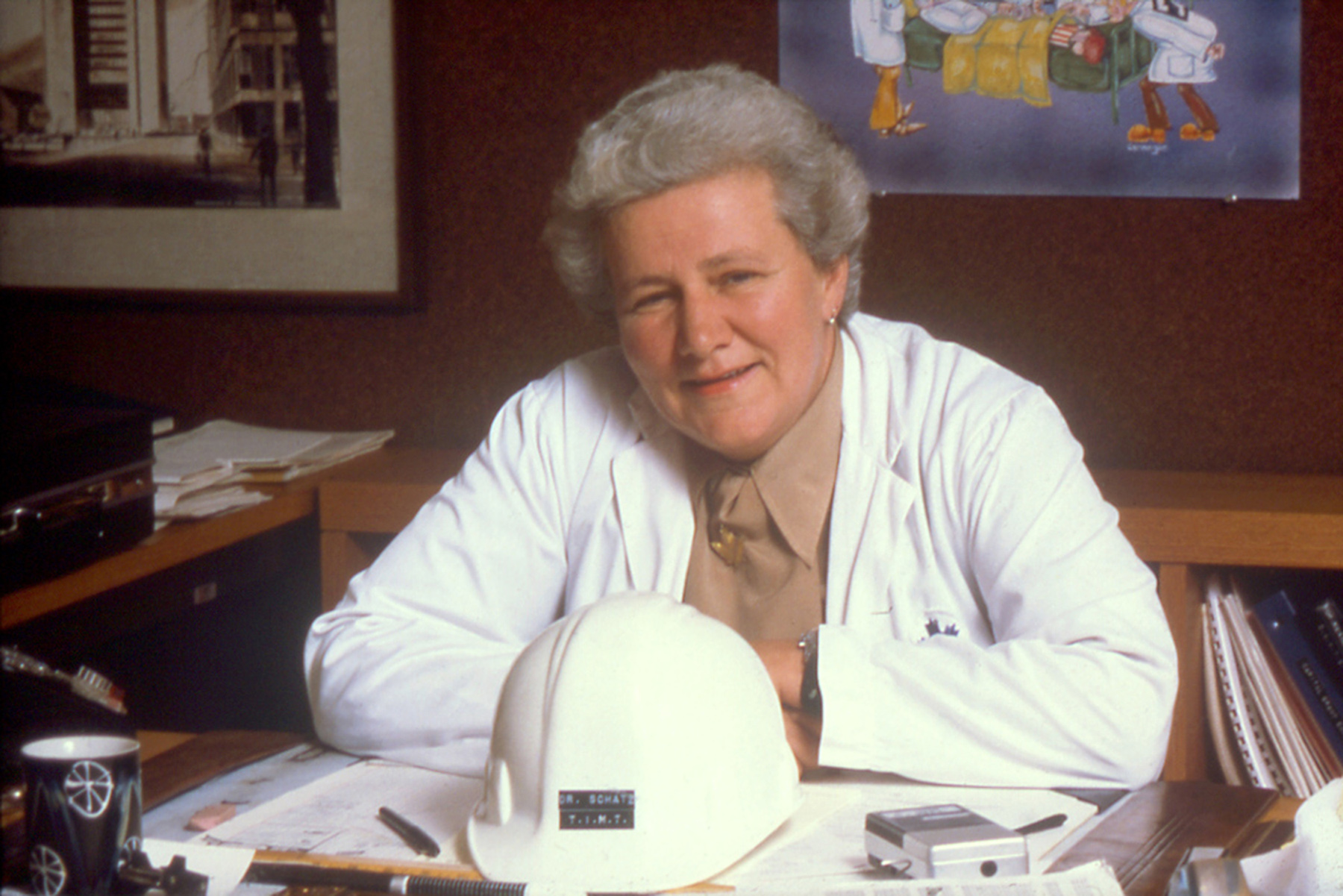 A person in a white lab coat sits at a desk with papers and a white hard hat in an office setting.