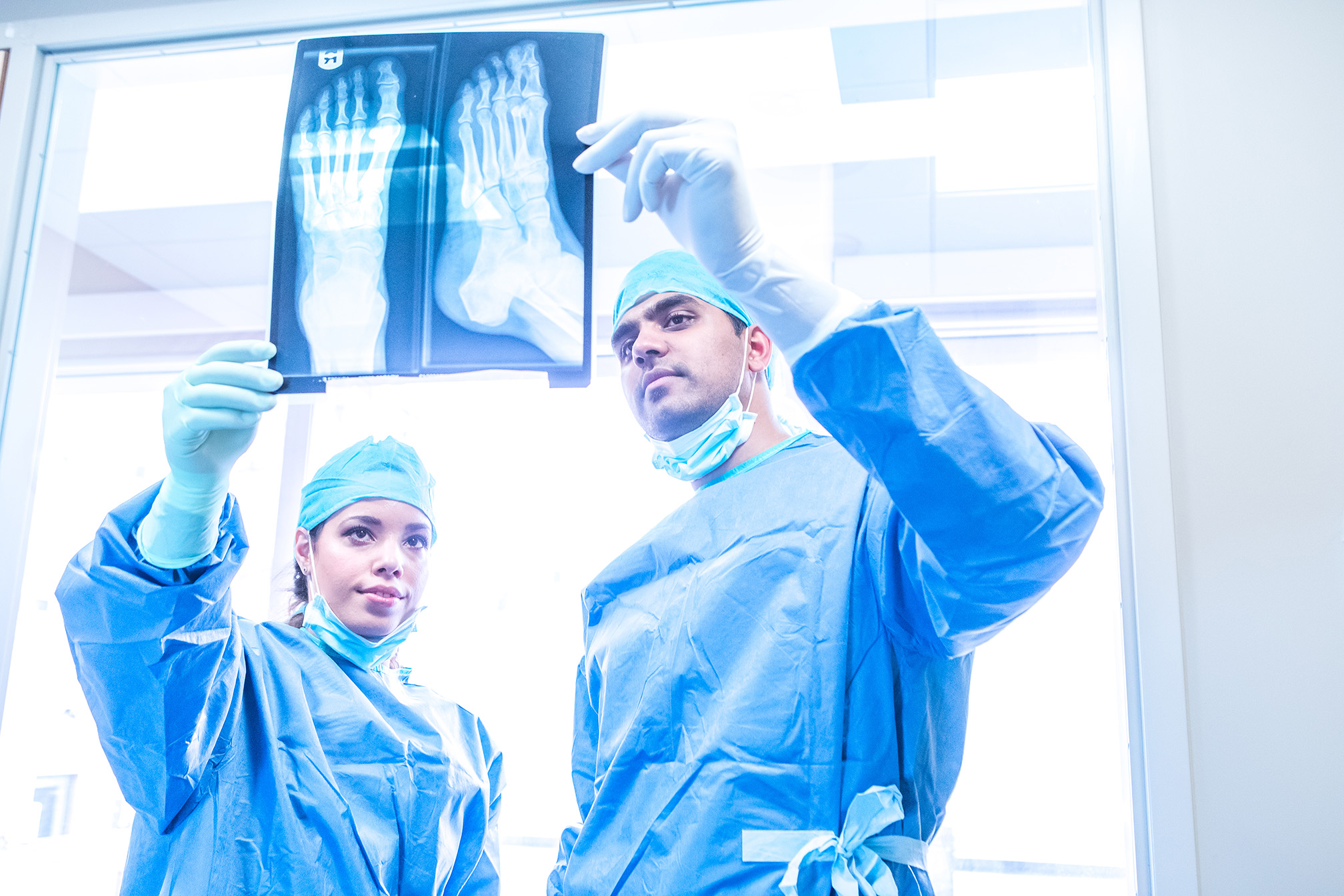 Two medical professionals in scrubs examine an X-ray image of feet in a brightly lit hospital room.