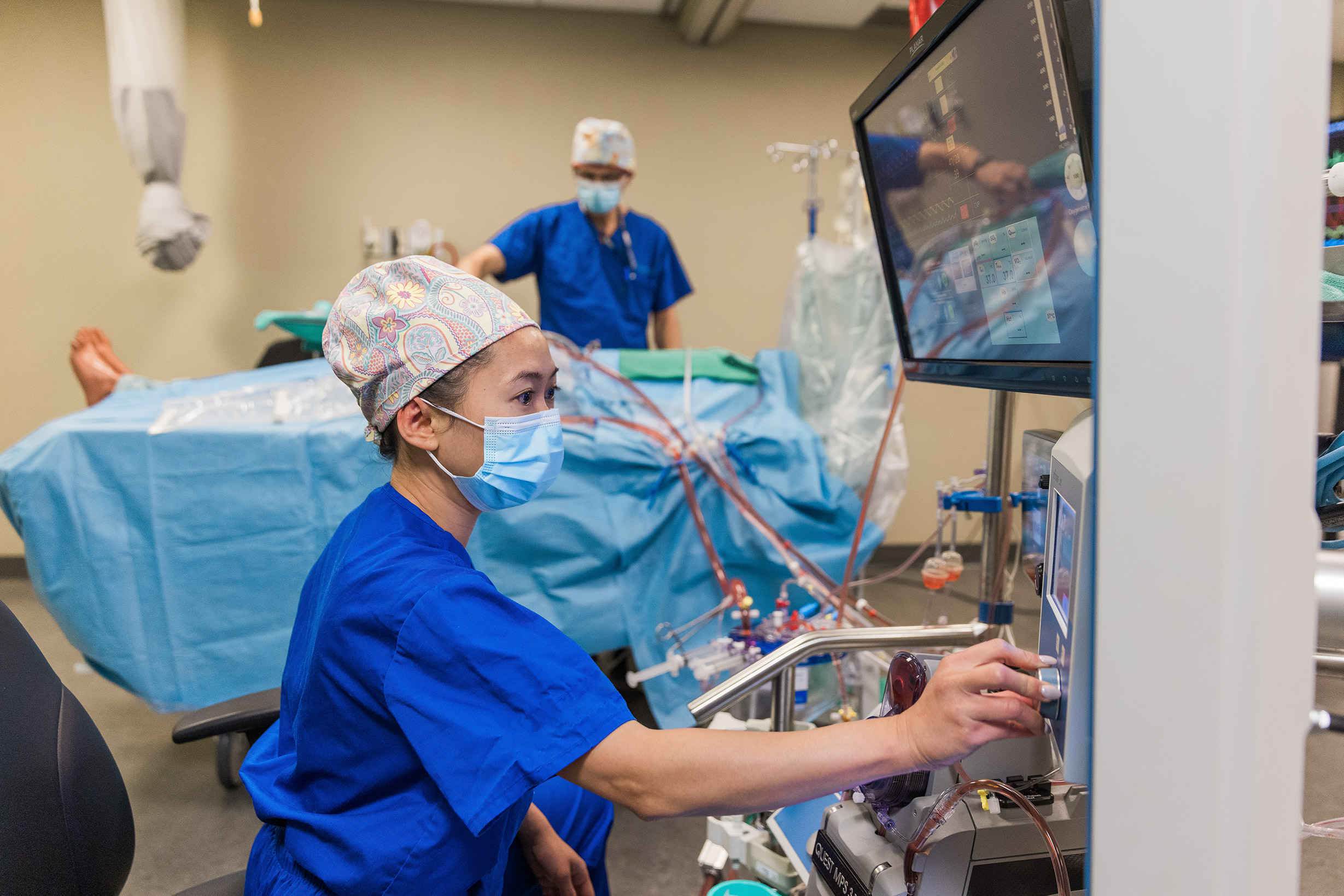 A medical professional operates equipment beside a patient in surgery while another person works in the background.