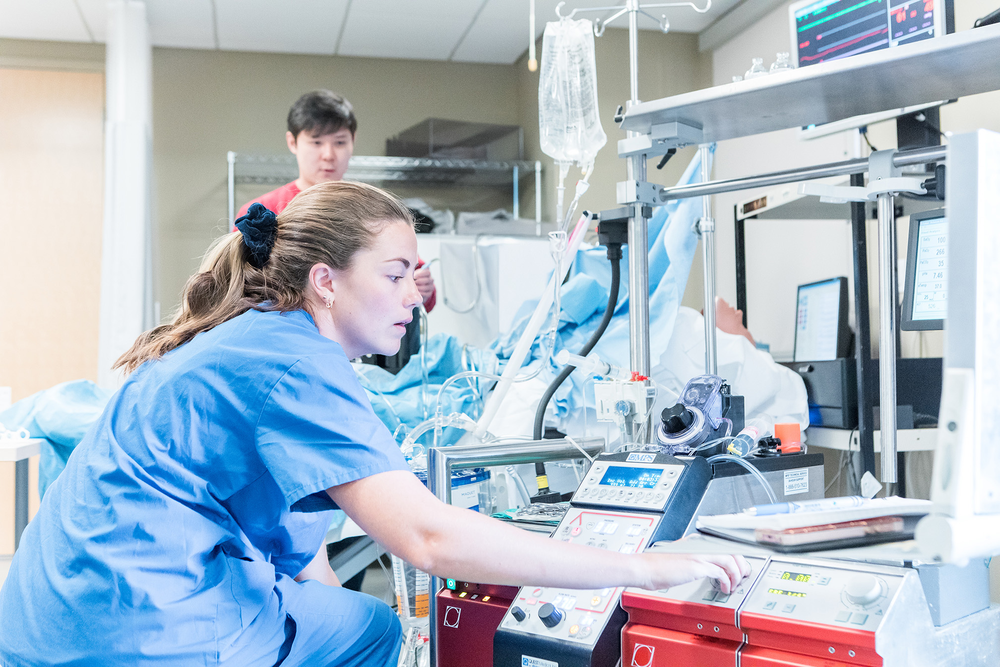 A healthcare professional in blue scrubs adjusts medical equipment in a hospital room with another person in the background.