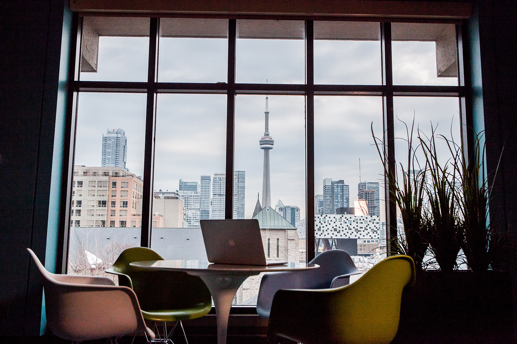 A laptop on a round table with chairs, set by a large window overlooking the Toronto skyline and CN Tower.