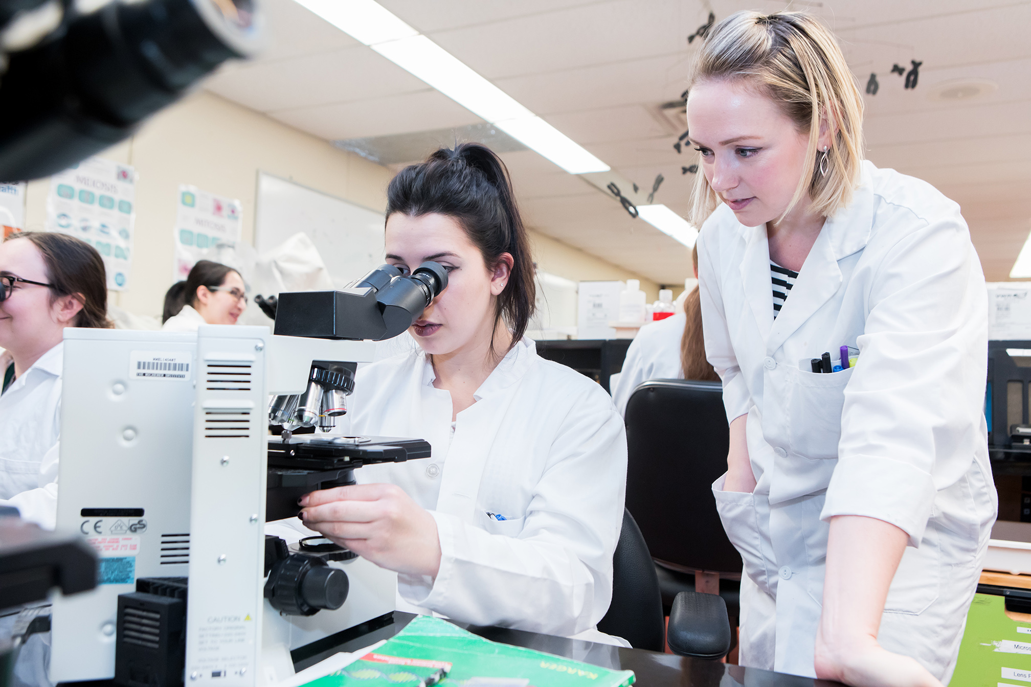 Two women in lab coats work with a microscope in a laboratory, one observing a slide while the other looks on.