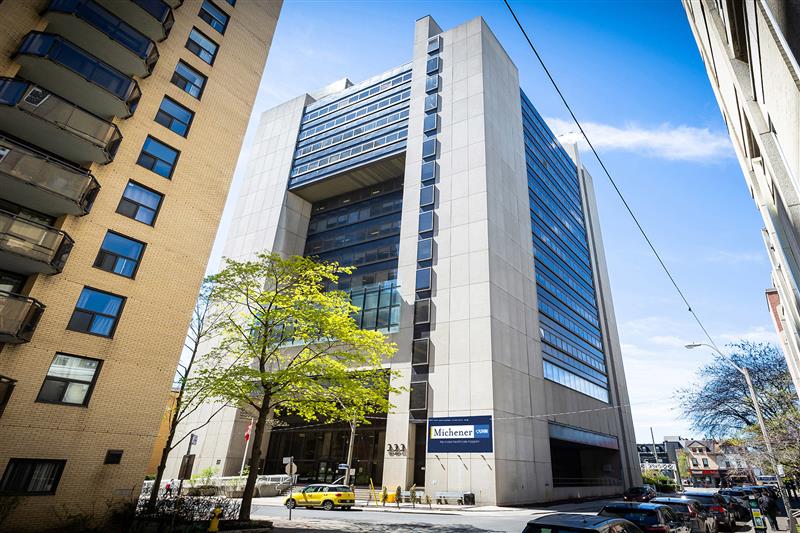 A tall, modern building with a Michener sign on its exterior, surrounded by other buildings and a few cars on the street.