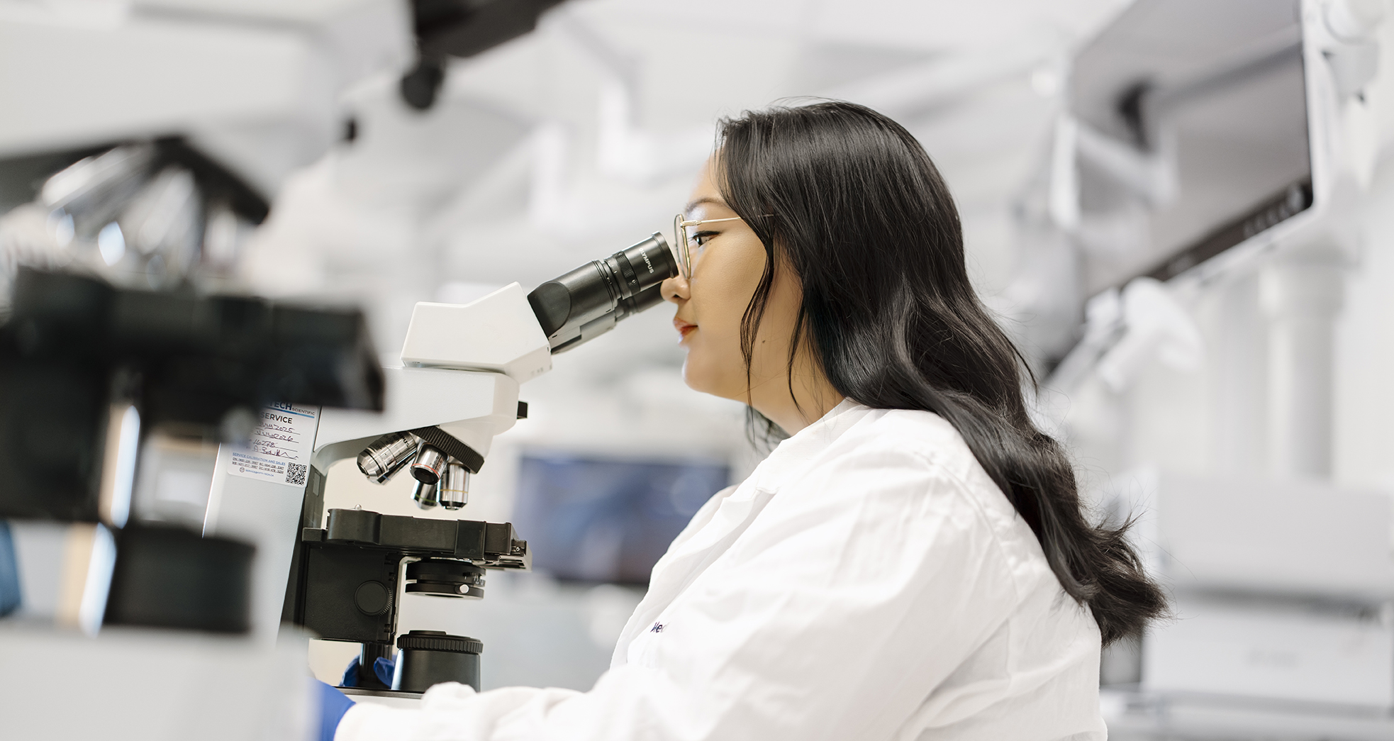 A scientist in a white lab coat observes samples through a microscope in a laboratory setting.