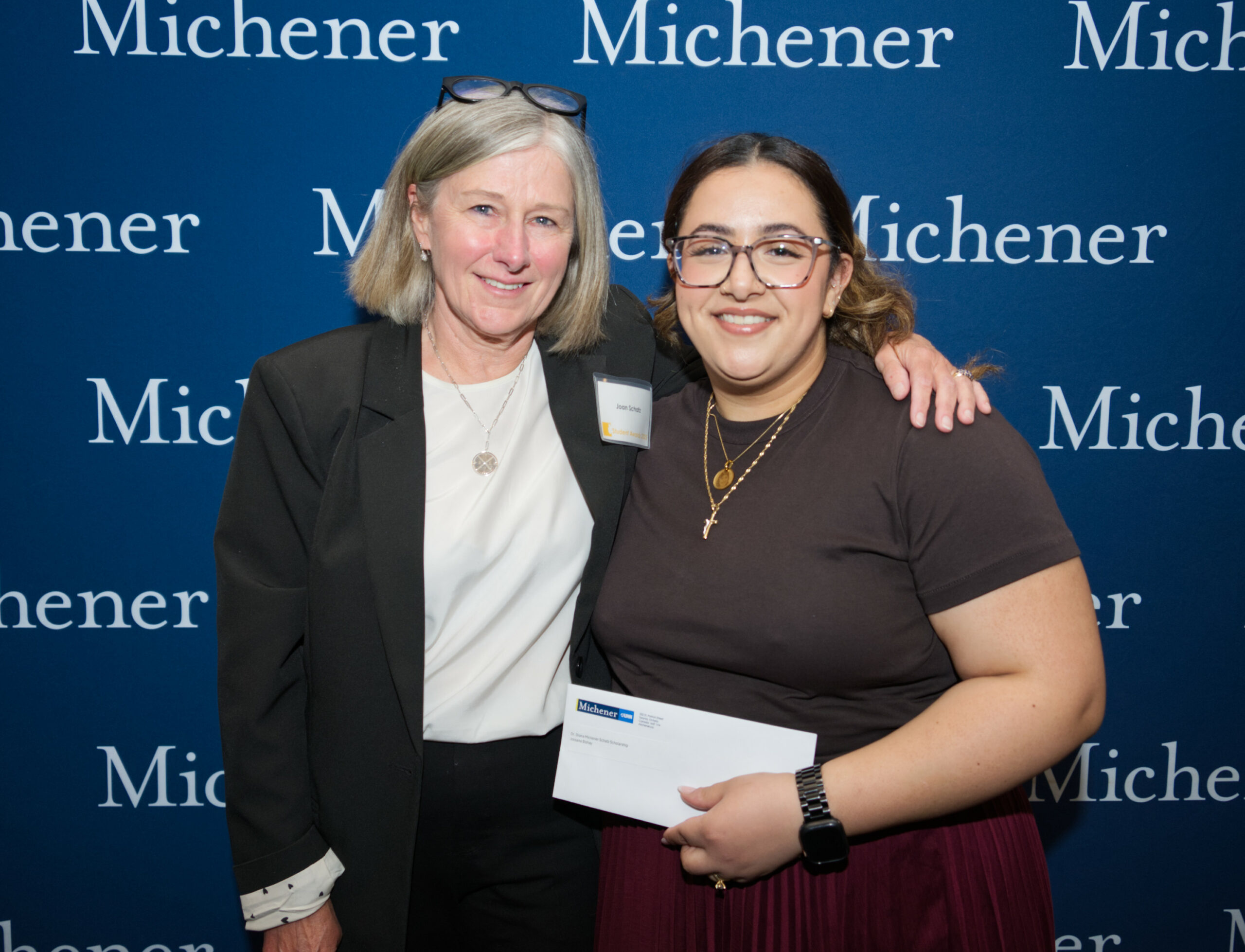 Two women pose together in front of a blue Michener backdrop, one holding an envelope and both smiling at the camera.
