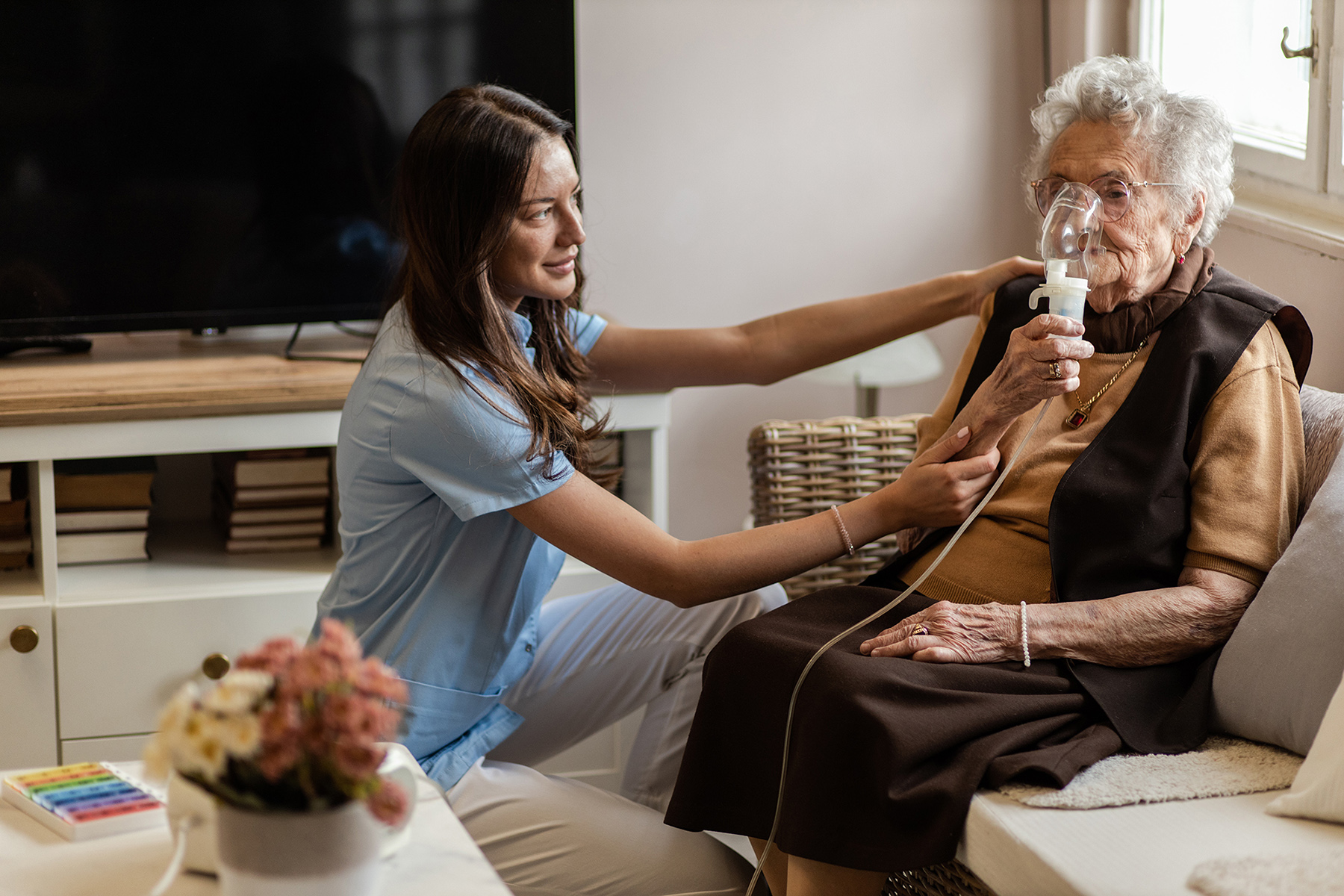 A nurse helps an elderly woman use a nebulizer while sitting together in a living room.