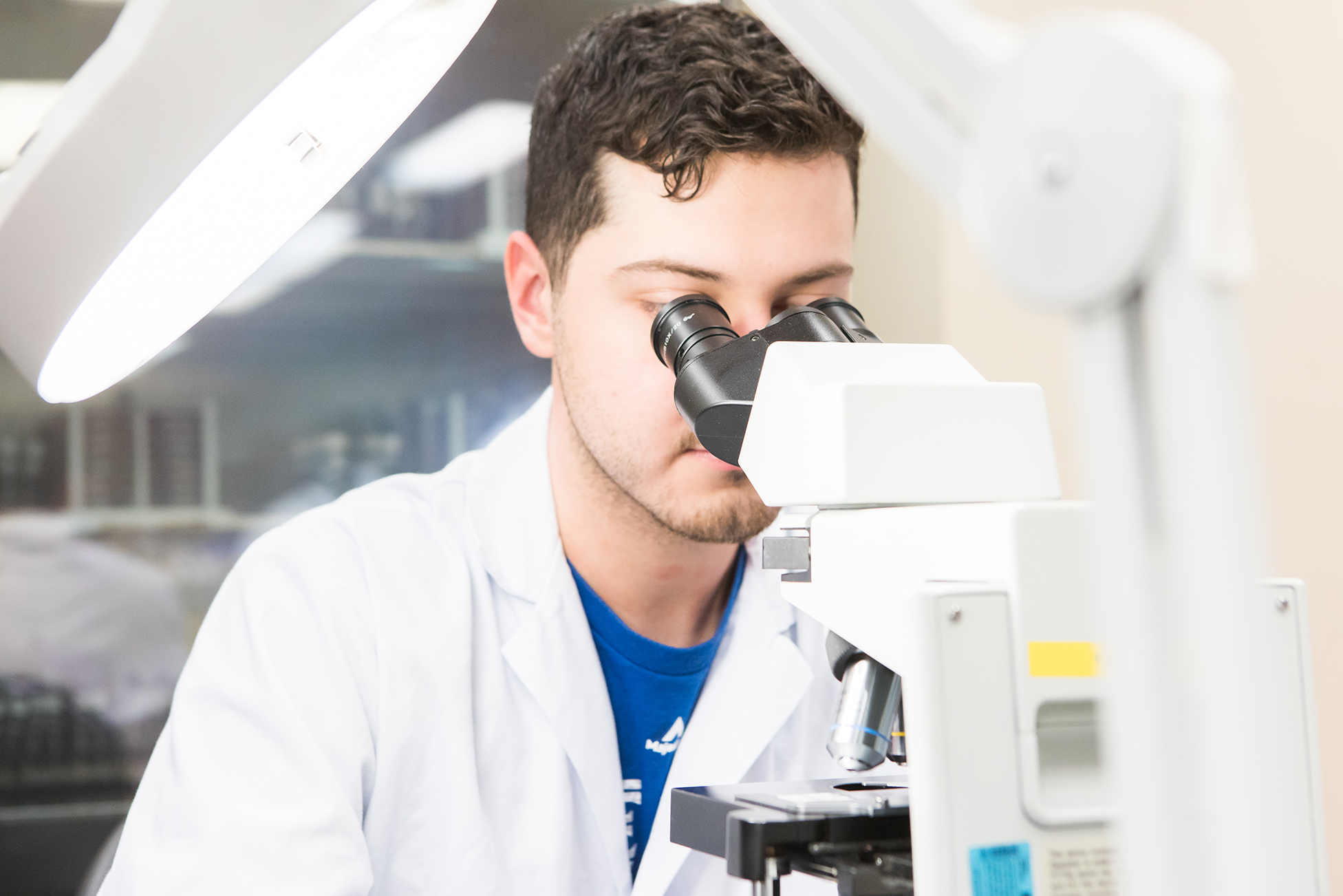A scientist in a lab coat looks into a microscope under bright lighting in a laboratory setting.