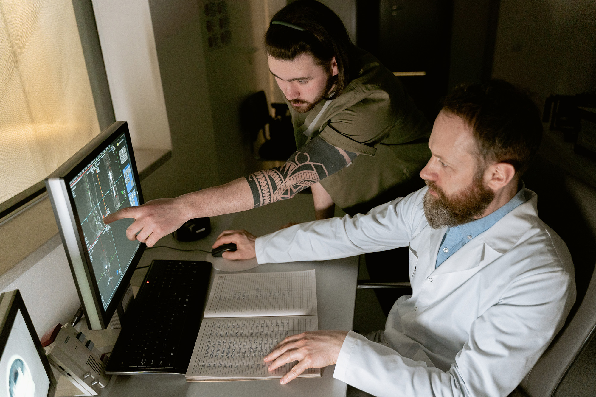 A man in a lab coat sits at a desk, while another man points at data displayed on a computer monitor.