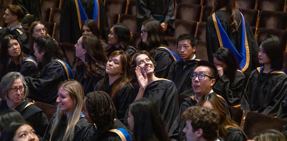 Graduates in black robes sit in an auditorium; one woman in the center smiles and waves at the camera.