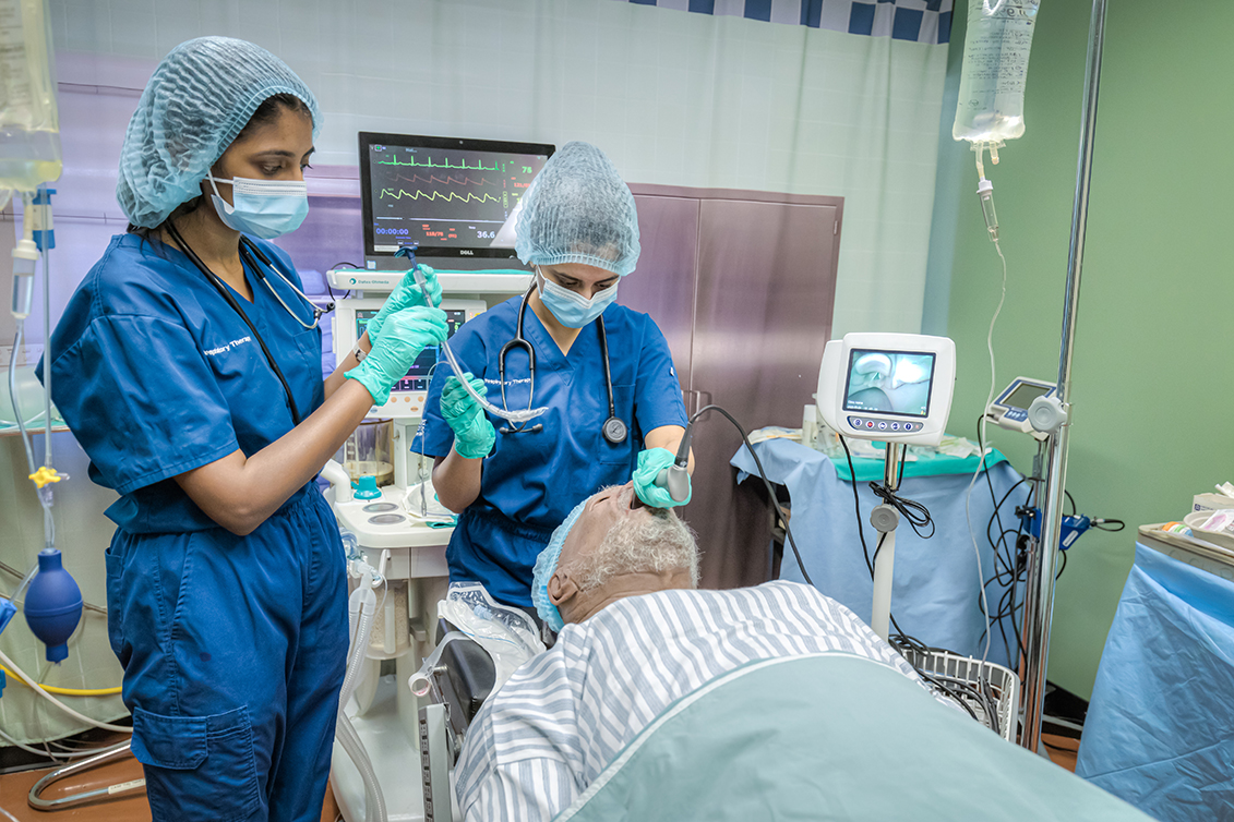 Two medical professionals prepare an older patient for a procedure in a hospital room with monitoring equipment visible.