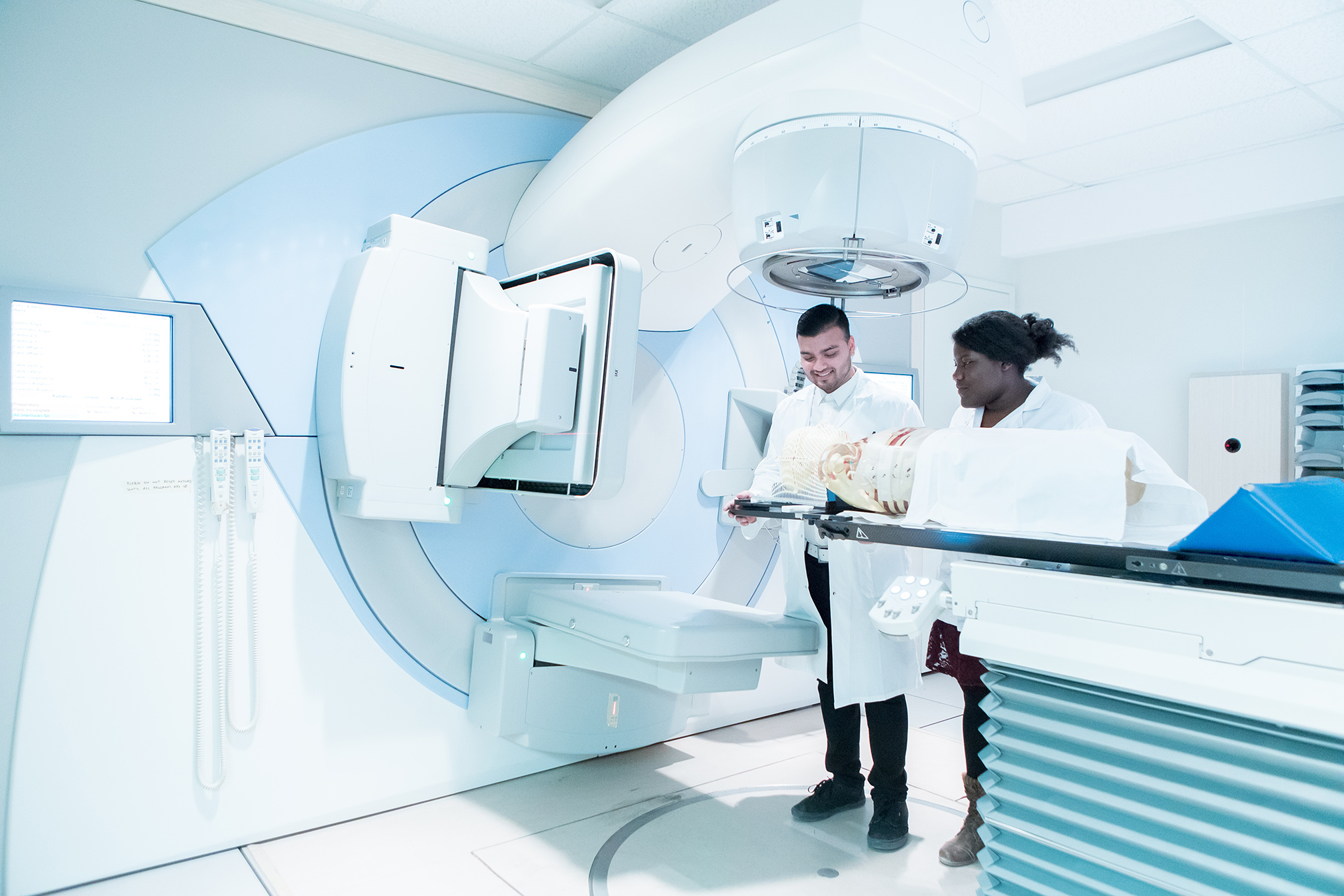 Two medical professionals prepare a patient for a procedure using advanced radiotherapy equipment in a hospital room.