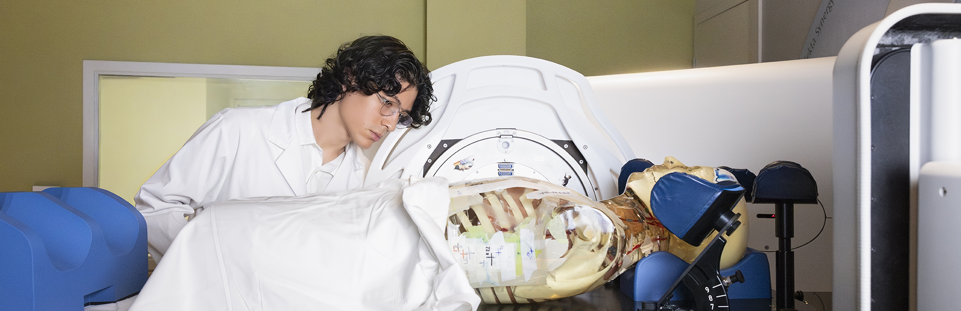 A person in a lab coat examines a medical mannequin on a scanning machine in a clinical setting.