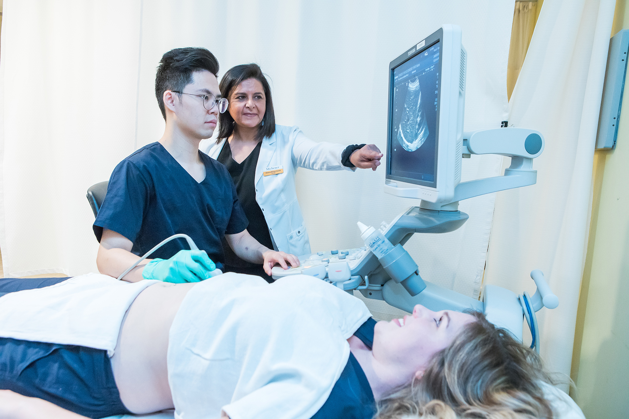 A medical professional performs an abdominal ultrasound on a patient as another clinician points to the monitor.