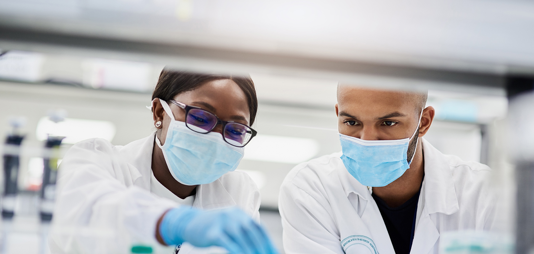 Two scientists wearing lab coats and face masks work together in a laboratory, examining samples on the table.