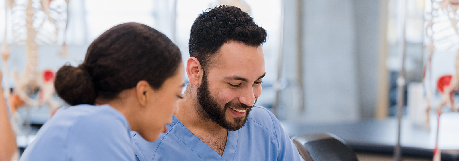 Two medical students in blue scrubs sit together, studying and discussing in a classroom with anatomical models.