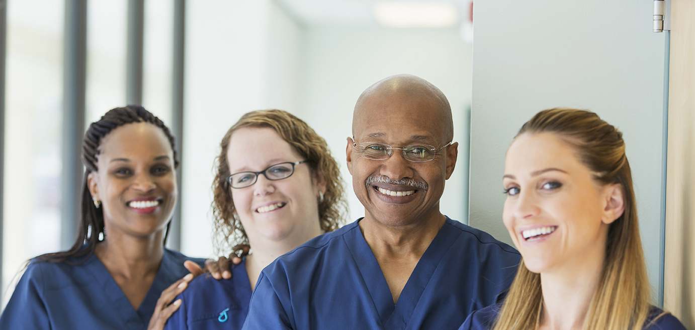 Four healthcare professionals in blue scrubs standing together and smiling in a well-lit hallway.