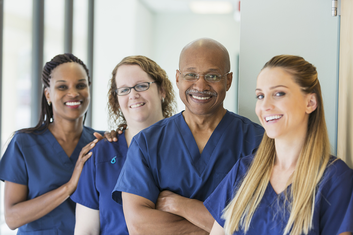 Four healthcare professionals in blue scrubs stand together, smiling, in a medical facility hallway.