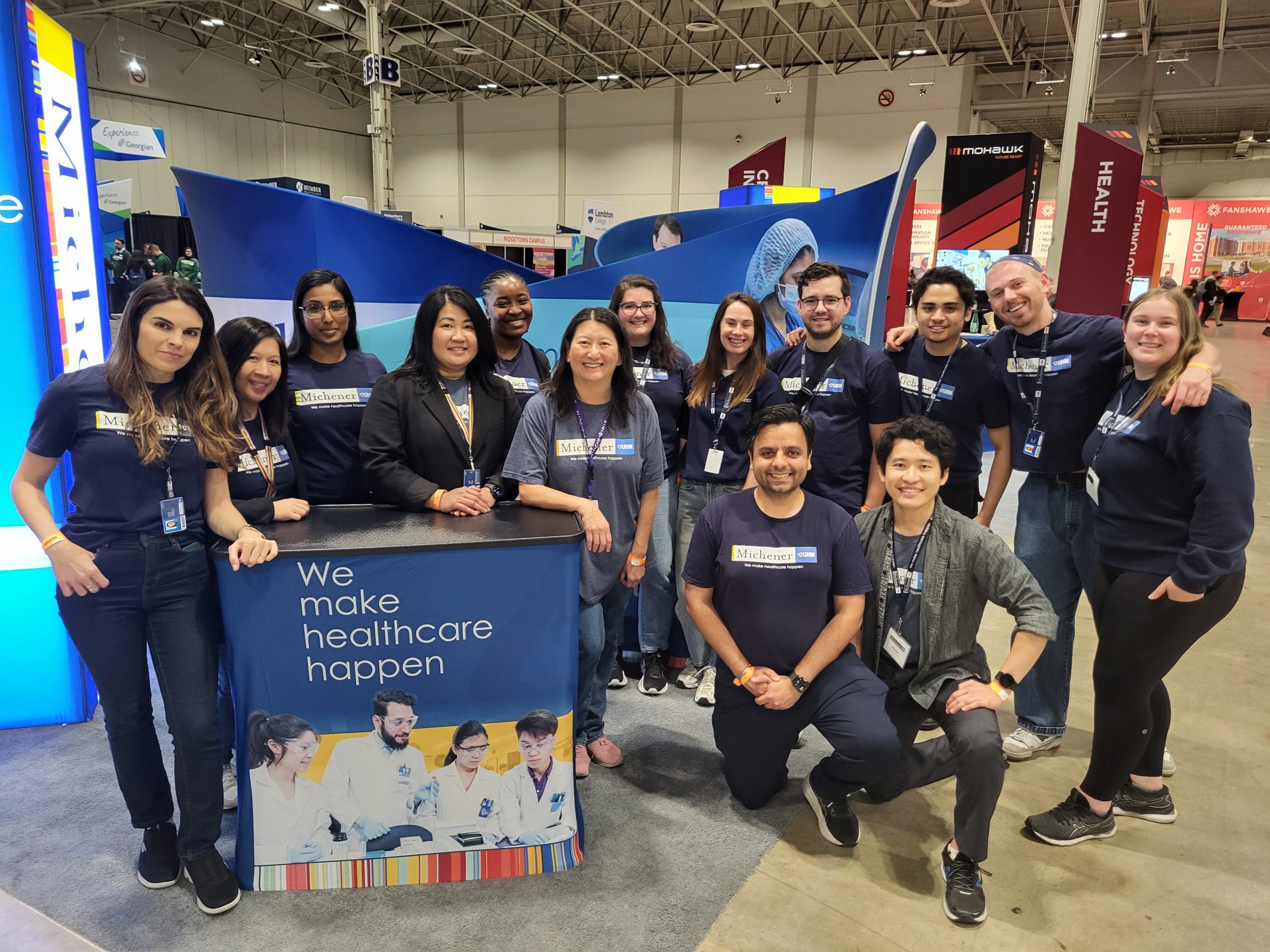 A group in Michener t-Shirts pose together at a healthcare event booth with a sign reading "We make healthcare happen.